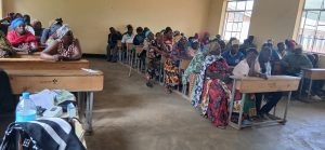 Classroom filled with adults seated at desks, listening to a health education session. Medical tools and supplies are visible on the front desk, symbolizing the outreach’s focus on screening and awareness.